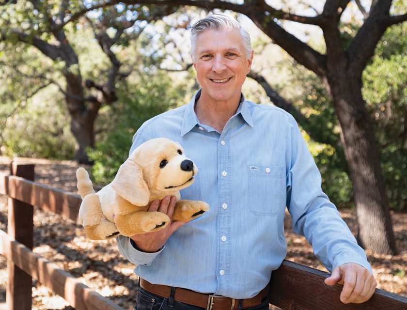 A smiling older man with gray hair stands outdoors holding a plush robotic dog. He wears a blue button-down shirt, and trees fill the background.